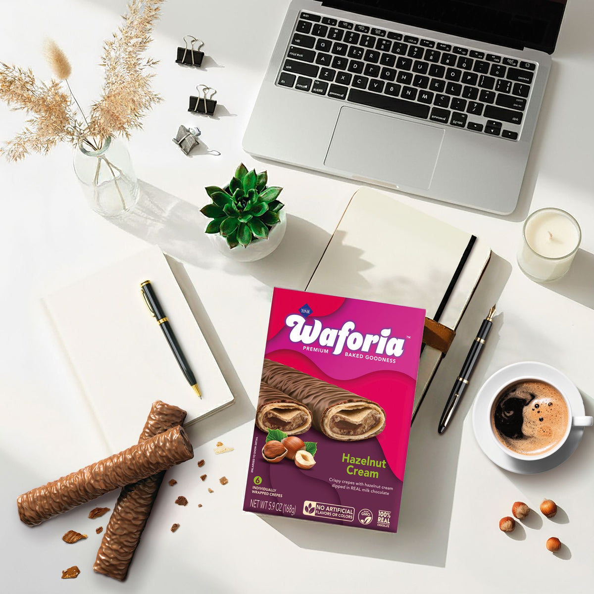 A flat-lay of a box of Waoria Fresh Baked Chocolates (Hazelnut Cream flavor) on a white desk surrounded by lifestyle props including a laptop, coffee, pens, and a succulent.