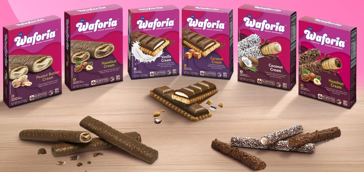 Six boxes of Waforia chocolate wafer bars in different flavors are lined up against a pink background, with several chocolate wafer bars and cookie crumbs displayed on a light wood surface in front.